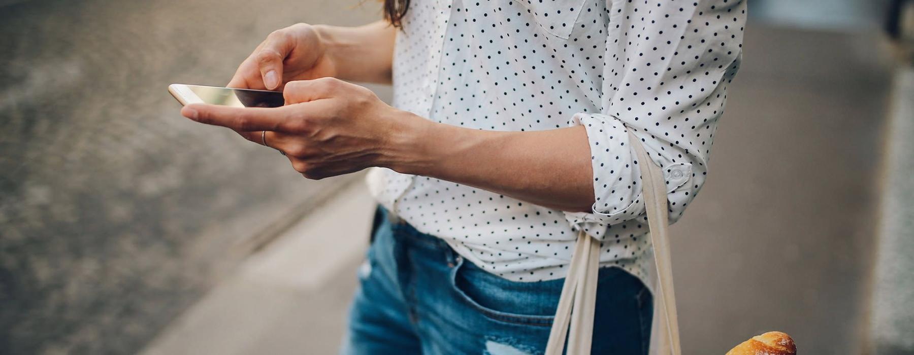 woman walks down the street and texts with a bag of groceries on her arm